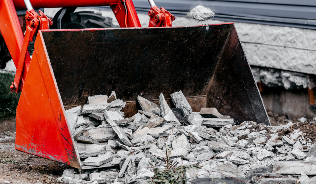 Red excavator bucket loading broken concrete slab chunks and rubble during concrete removal and demolition cleanup project