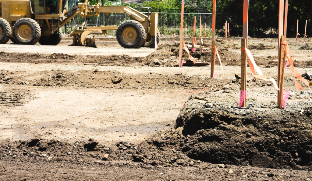 Motor grader performing rough grading on a Florida construction site with survey stakes and grade flags marking spot elevations across the disturbed soil surface